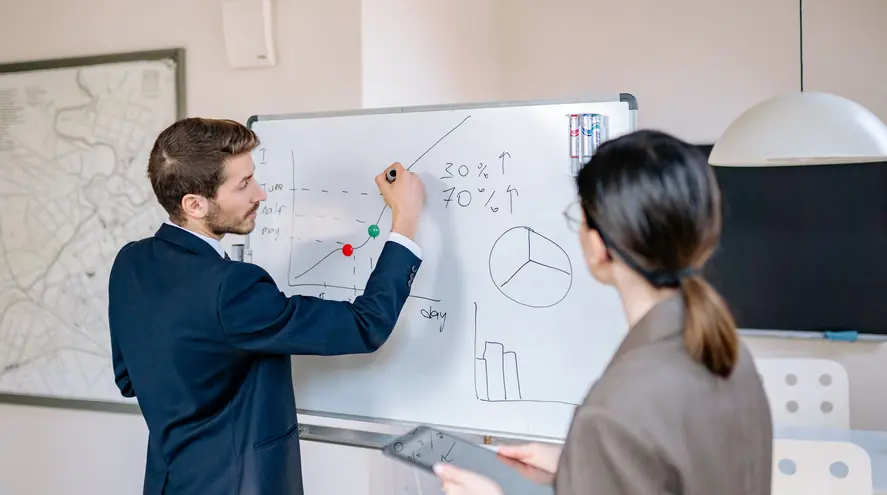 A man in a navy suit sketches a line graph on a whiteboard while a colleague holding a tablet watches