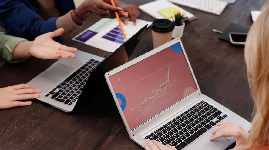Colleagues around a wooden meeting table with two open laptops and a printed bar chart